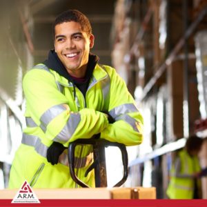 Image of smiling handsome young male warehouse worker in yellow jacket with flare stripes, leaning on the handle of a pallet jack loaded with boxes with warehouse in background for safety tips ARM action resource management