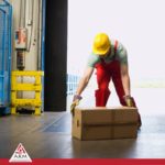 Image shows warehouse worker in red overalls and yellow safety cap and gloves lifting a box from the ground for safety tips ARM action resource management 