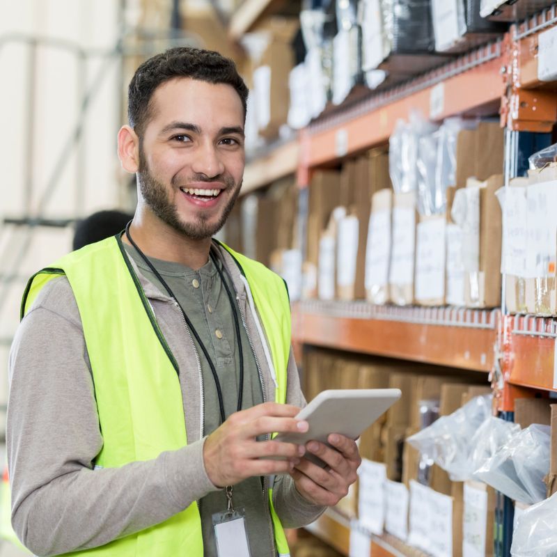 Image of smiling young bearded warehouse worker holding a tablet near product shelves for A.R.M. action resource management