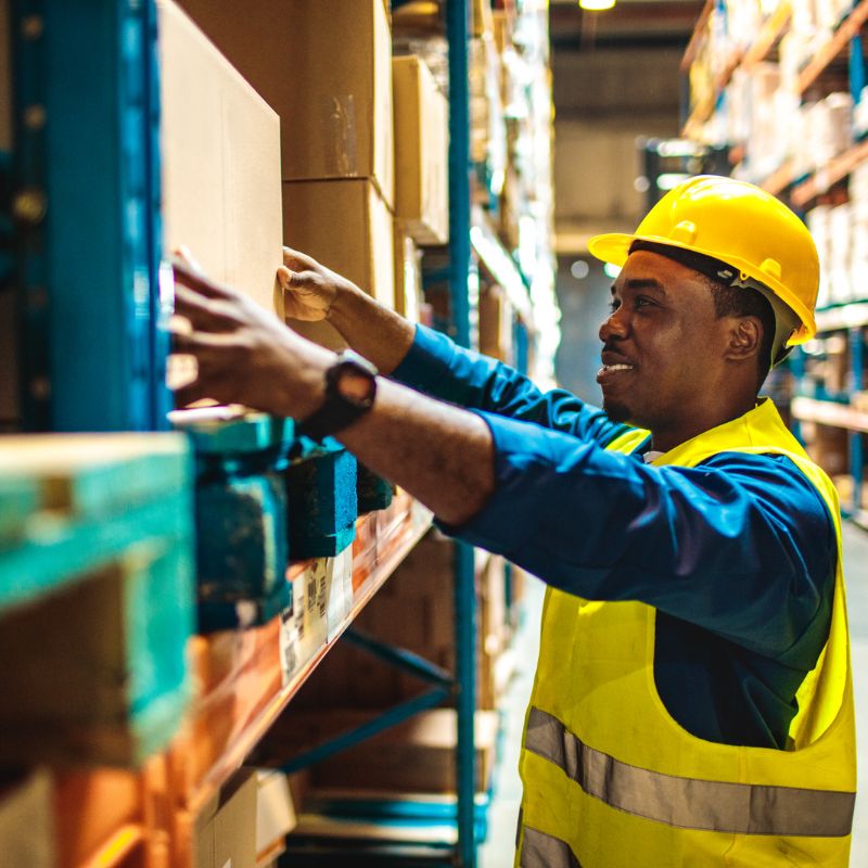 Image shows profile of smiling warehouse worker with pushed-up sleeves reaching for a box clothing safety short sleeves warehouse A.R.M. action resource management
