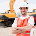 Construction worker in orange safety vest and white hard hat standing in front of machinery with their arms crossed and smiling. Outdoor worker safety. A.R.M. action resource manaegmenet