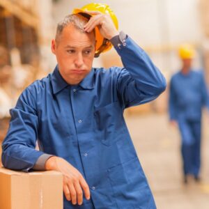 Image of warehouse worker man dressed in blue uniform leaning against boxes looking flustered or worried, with warehouse behind him A.R.M. action resource management