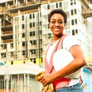 Image of woman construction worker with short natural hair, orange safety vest, holding gloves and hard hat, smiling with building behind. A.R.M. action resource management