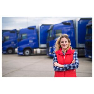 Image of young woman truck driver in red vest, arms crossed, wind in hair, smiling in front of a row of parked blue semi trucks. A.R.M. action resource management