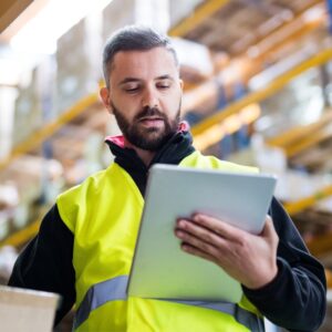 Image of bearded young warehouse worker wearing yellow safety vest over dark hoodie studying his handheld tablet. A.R.M. Action Resource Management Safety training