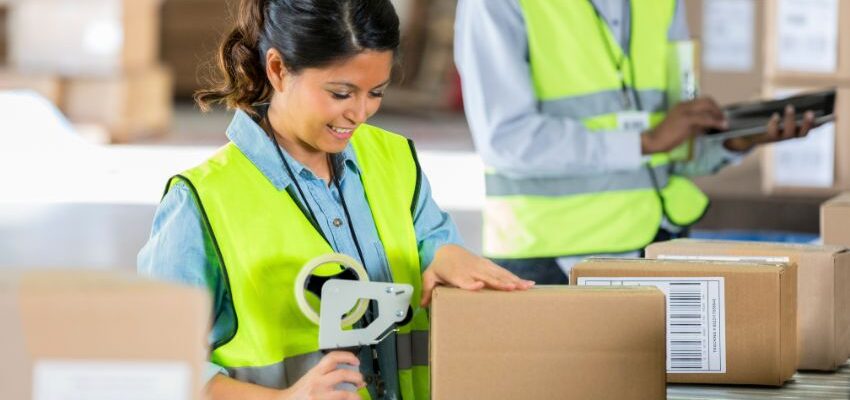 Image shows smiling young woman warehouse worker wearing a yellow safety vest and taping a box closed. General warehouse worker Fontana. A.R.M. Action Resource Management.