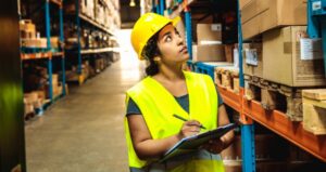 Image shows young woman warehouse worker wearing yellow hard hat and safety vest, holding a clipboard and pen while examining stock on shelves. general warehouse worker. Action Resource Management. A.R.M.