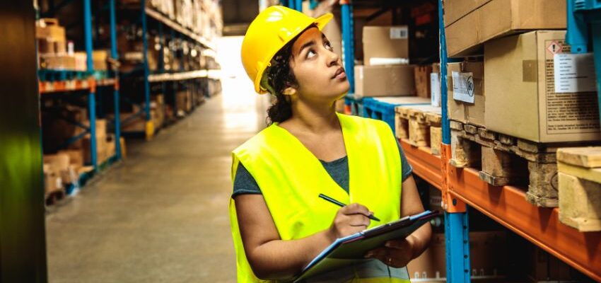 Image shows young woman warehouse worker wearing yellow hard hat and safety vest, holding a clipboard and pen while examining stock on shelves. general warehouse worker. Action Resource Management. A.R.M.
