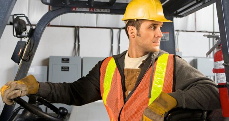 Image of young man forklift driver wearing yellow hard hat and orange, yellow-striped safety vest looking over his shoulder as he backs up in forklift. Back to basics: forklift safety. A.R.M. Action Resource Management. safety training and services.
