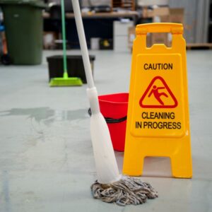 Image of a yellow “caution” sign next to a mop, bucket, and water on floor of a warehouse. A.R.M. action resource management