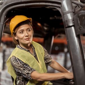 Image of young woman forklift operator wearing yellow hard hat and yellow safety vest looking ahead as she drives. A.R.M. Action Resource Management. safety training and services.