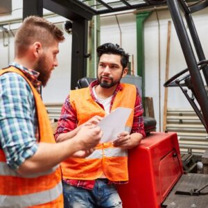 Image of two young warehouse workers, both in orange safety vests, one leaning on a forklift, the other, a safety trainer, reviews notes. A.R.M. Action Resource Management