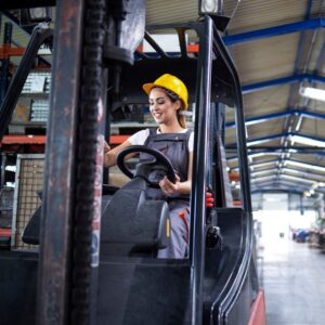 Image of a young forklift operator wearing blue overalls and a yellow hard hat, smiling as she drives the machine in a warehouse. A.R.M. Action Resource Management.