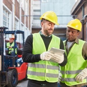 The image shows two men reviewing information on a tablet, both wearing gloves, long sleeves, hard hats, and safety vests. There is a person operating a forklift in the left-side background. Winter safety tips for outdoor workers. Action Resource Management A.R.M. Safety Training