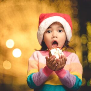 Image of a child wearing a Santa hat and colorful holiday sweater being surprised as she holds a Christmas gift. A.R.M.