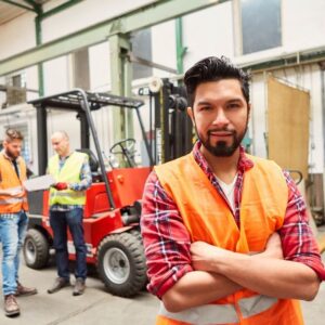 A warehouse worker wearing a white tee shirt, red flannel shirt, and safety vest stands, arms crossed, looking at camera, while two other workers confer over a clipboard, standing beside a forklift.