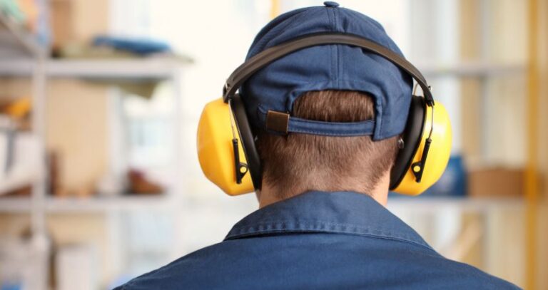 An image from behind of a warehouse worker wearing a blue shirt and cap, with yellow PPE ear protection.