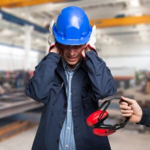 An image of a warehouse worker wearing a blue shirt, blue jacket, blue helmet and white gloves, holding his hands over his ears because it's too loud. Someone off camera is handing him ear protection PPE.
