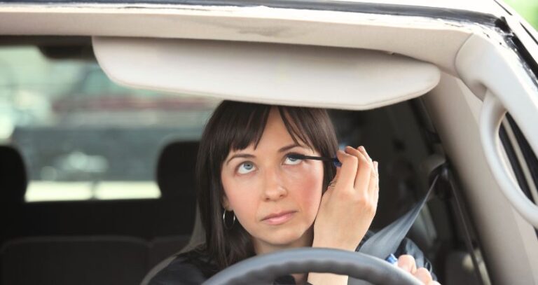 An image of a young woman applying mascara while driving her car. Distracted Driving Month - April