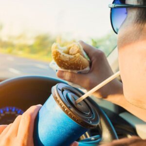 An image of a person driving their car while sipping a to-go drink from a straw in one hand and holding a sandwich and the steering wheel in the other. distracted driving