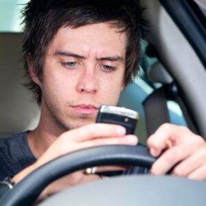 A young man who is driving his car with his brow furrowed as he studies his smartphone.