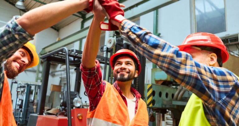 A group of three warehouse workers do a high five together, smiling about warehouse Safety in Seven