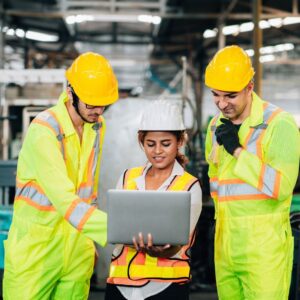 A woman warehouse manager is standing with her laptop, between two male warehouse workers dressed in yellow safety suits and hardhats, as they all look at something on the computer.