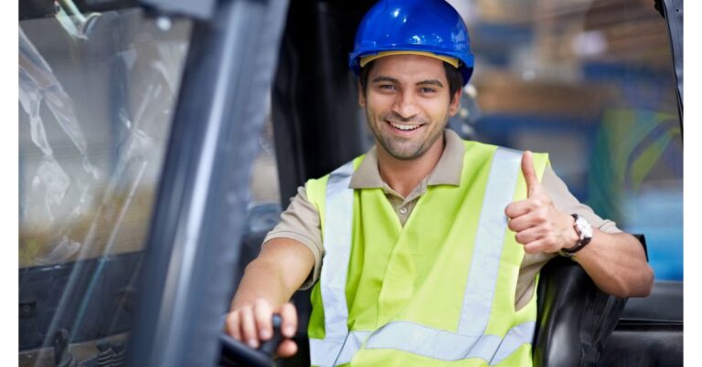 An image of a young forklift operator wearing a blue hard hat and yellow safety vest as he smiles and gives a thumbs up with his left hand.