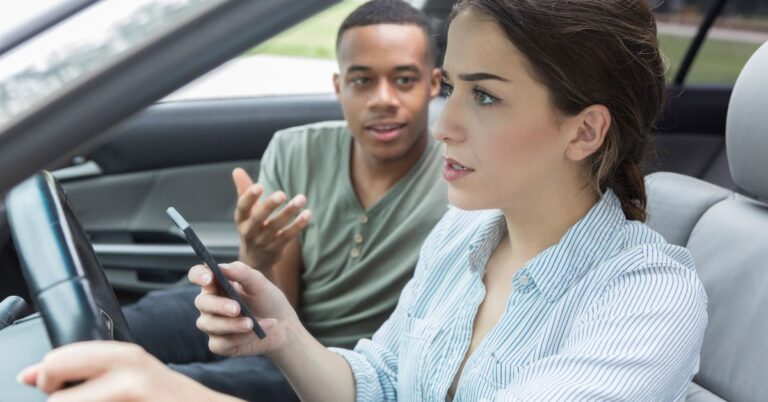 April is Distracted Driving Awareness Month: Stay Focused on the Road. A woman driver using her phone with one hand while her companion tries to argue.